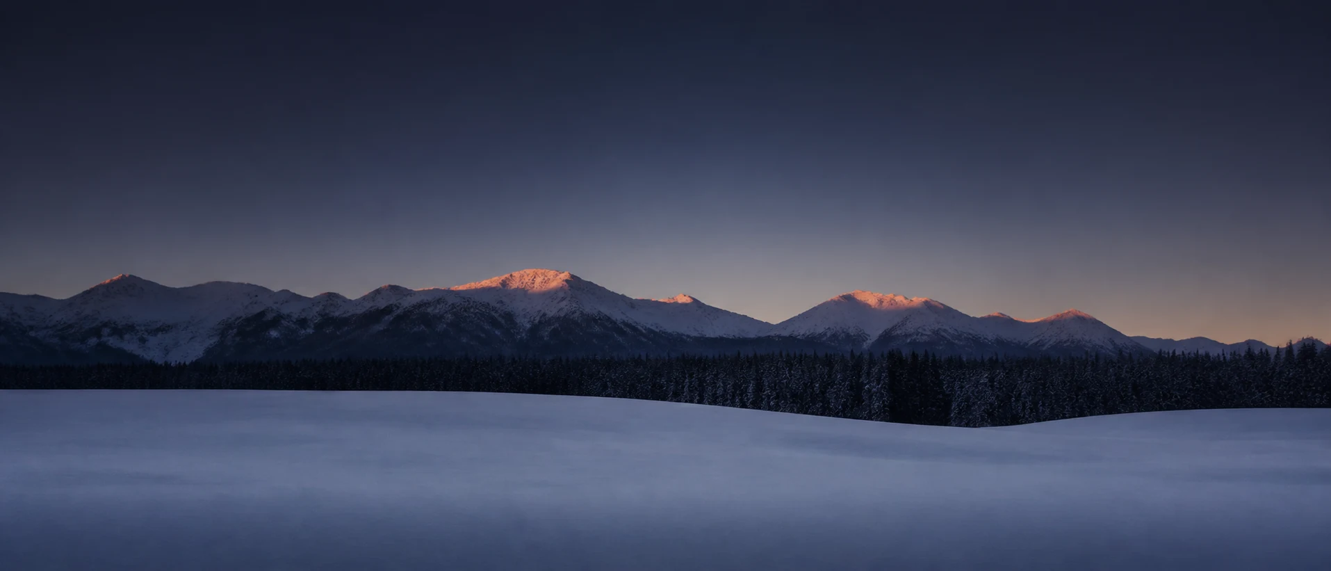 An open meadow in deep blue shadow with the peaks beyond still holding the last light